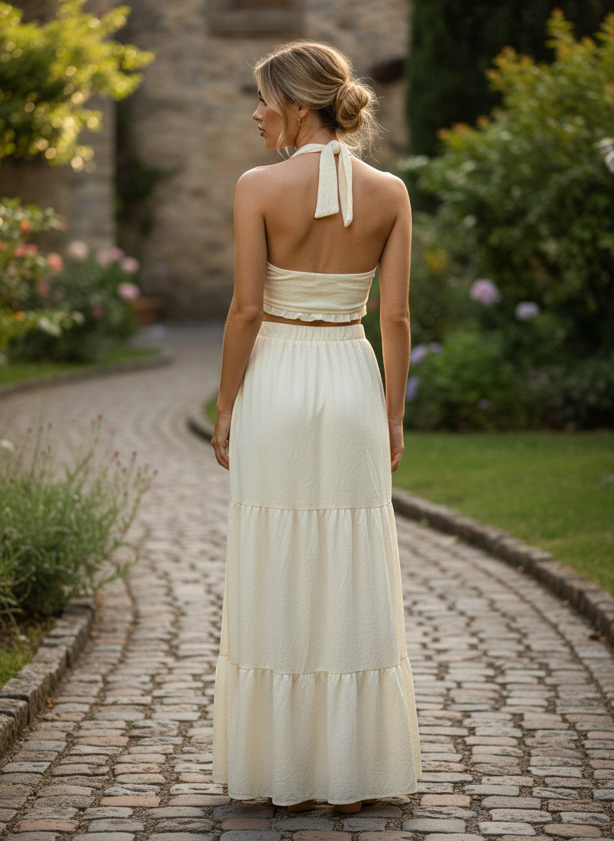 URHER Nadia cream two-piece dress: halter neck crop top with twist knot and tiered maxi skirt. Model from behind.